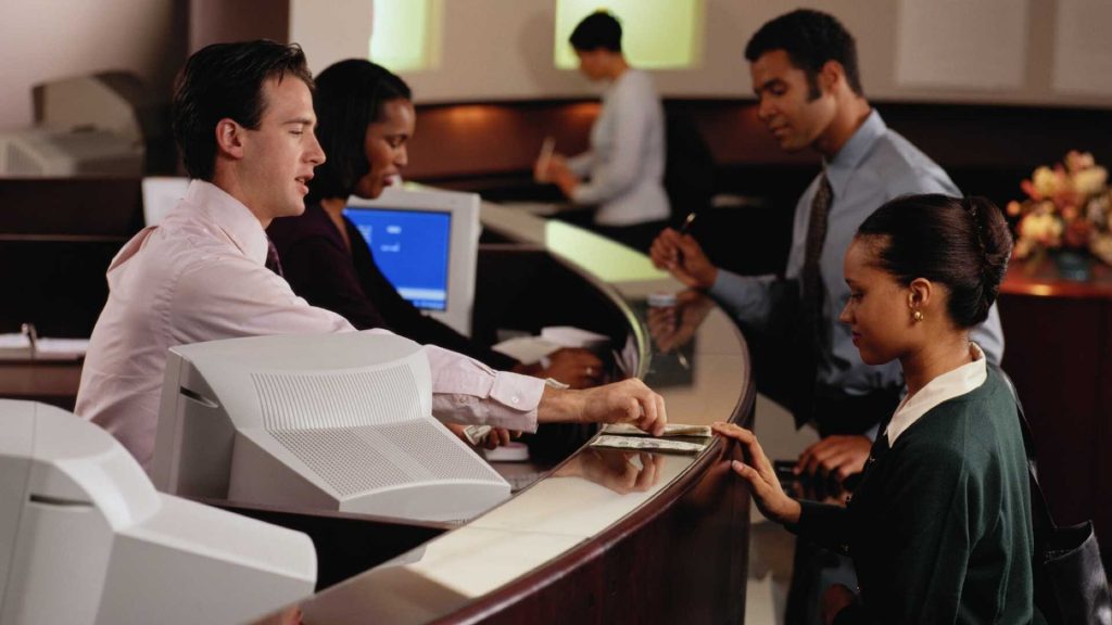  A bank teller is seen having a conversation with a customer while two other customers are waiting in line behind her.